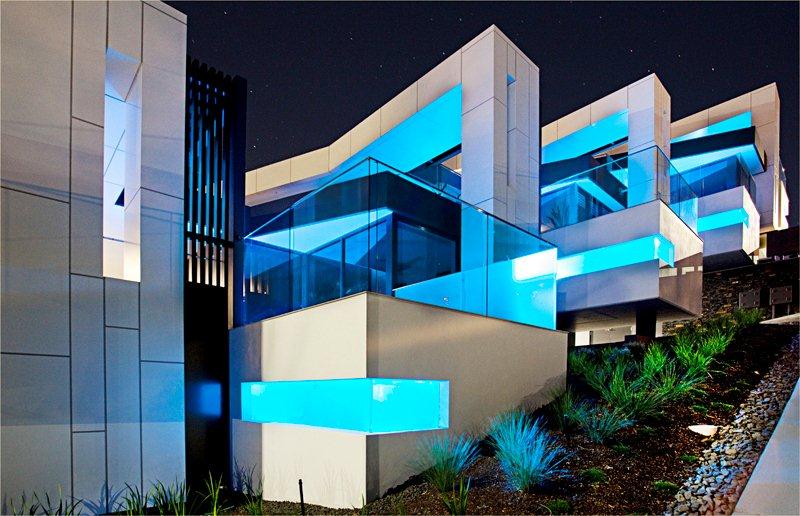 A luxurious residential development of four residences illuminated at night. The residences feature pools extending off the sides, adorned with blue lighting that shines upward onto the pristine white finished facades.