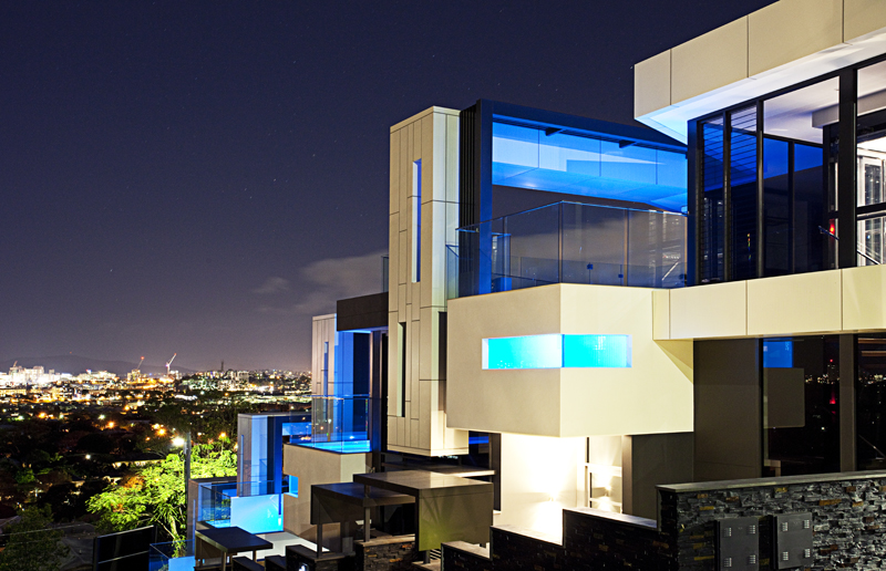 Main Avenue Bulimba development of four residences illuminated at night. The residences feature pools extending off the sides, adorned with blue lighting that shines upward onto the pristine white finished facades.