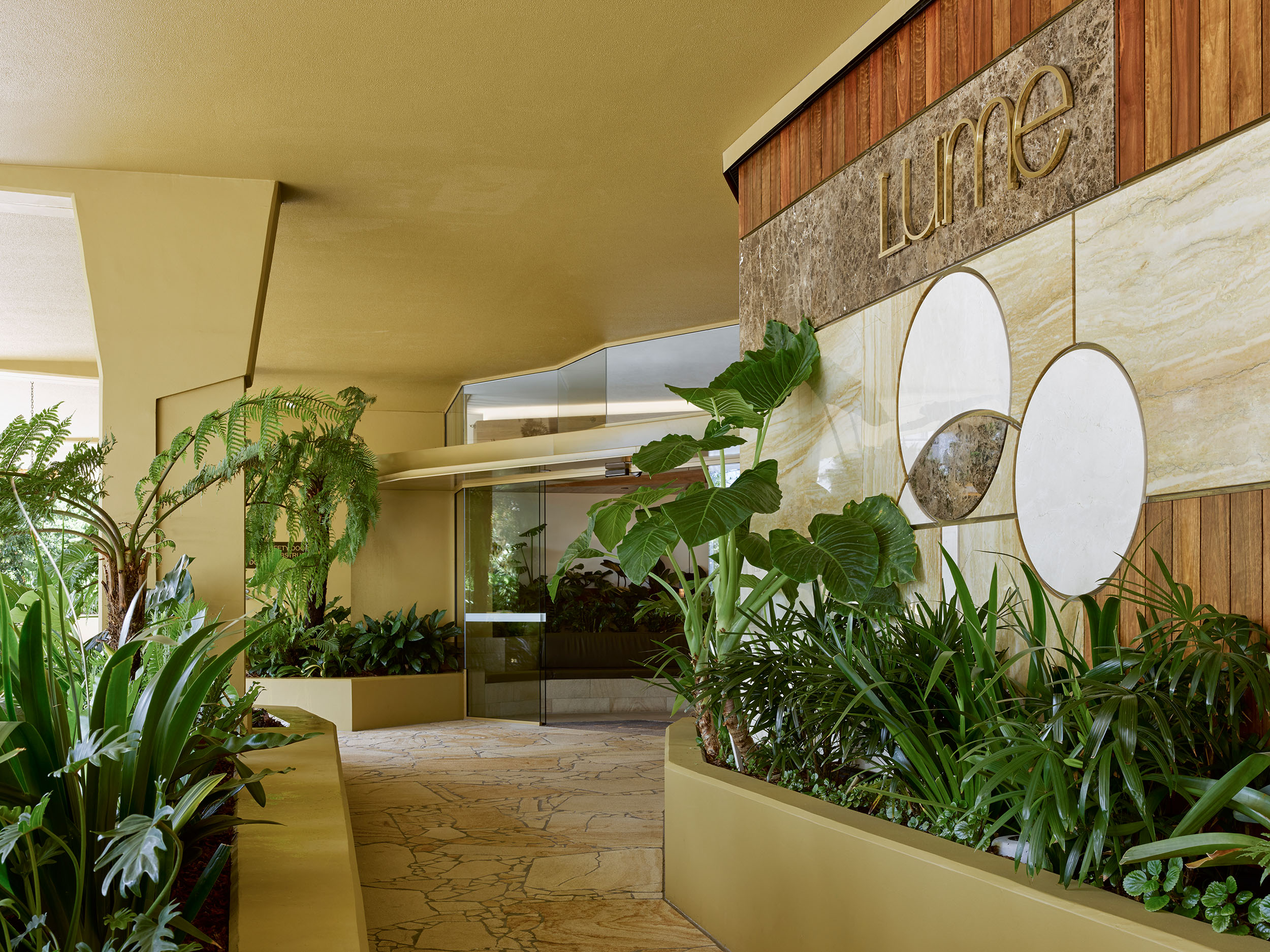 Entrance to a high-class apartment building, offering a picturesque view through a pathway created by two large planters filled with lush tropical plants. The entrance features gold accents, timber finishes, and a natural stone floor, exuding a luxurious and inviting ambiance.