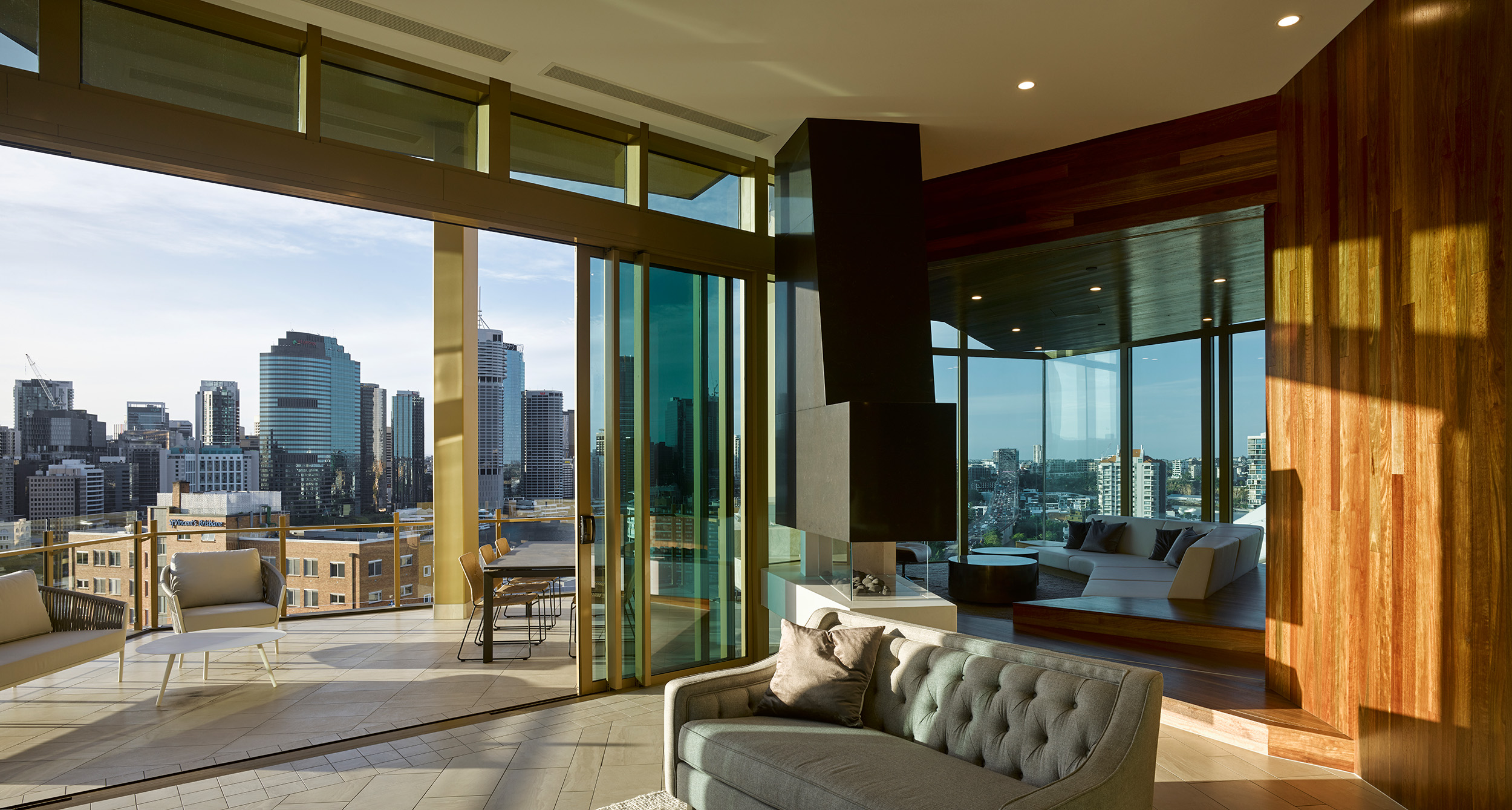 Stunning view from a penthouse apartment, showcasing the lounge area with premium furniture, tile floors, gold accents, and a beautiful timber wall. The space is surrounded by expansive glazing, flooding the interior with abundant natural light.