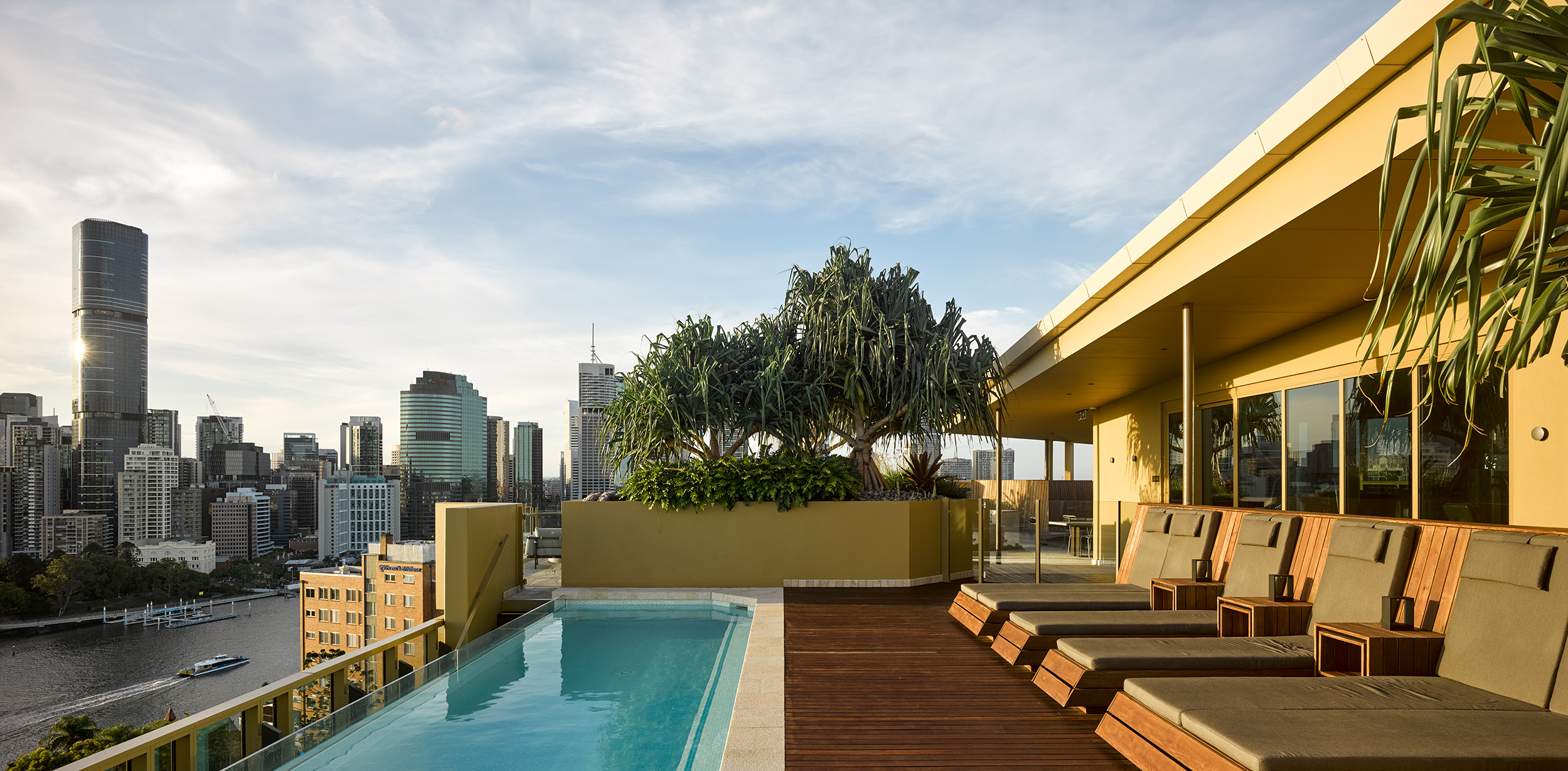 Breathtaking sunset view from a high-rise rooftop infinity pool, offering a stunning panorama of the Brisbane city skyline. The space features dark timber accents and luxurious gold color finishes. Abundant greenery and planting add a touch of natural beauty, creating a premium and serene atmosphere.